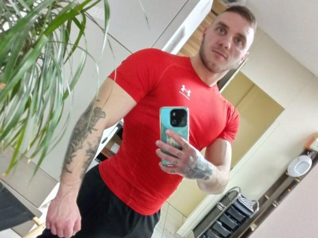 JordanDiary with a muscular build, short brown hair, tattoos on arms, wearing a tight red Under Armour shirt and black pants, taking a selfie with a teal phone.