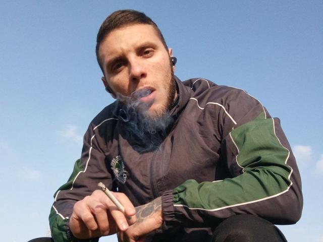 JordanDiary with short brown hair and beard, wearing a green and gray track jacket, smoking a cigarette with blue smoke, against a clear blue sky.