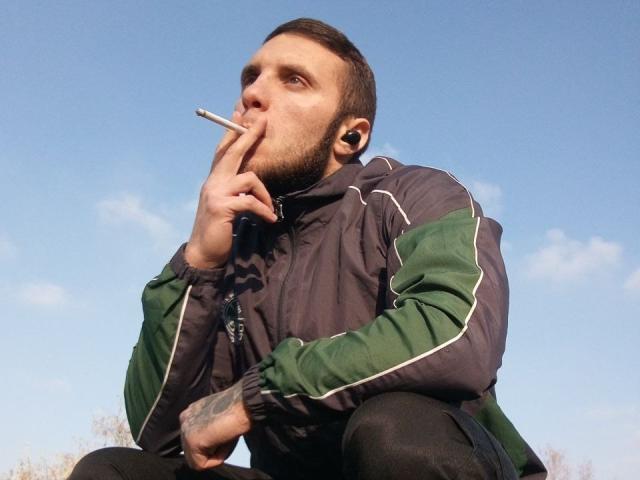 JordanDiary, squatting, puffs cigarette, green and gray jacket, black pants, tattoos on right hand, short brown hair, beard, blue sky, earbuds in, serious expression, daytime.