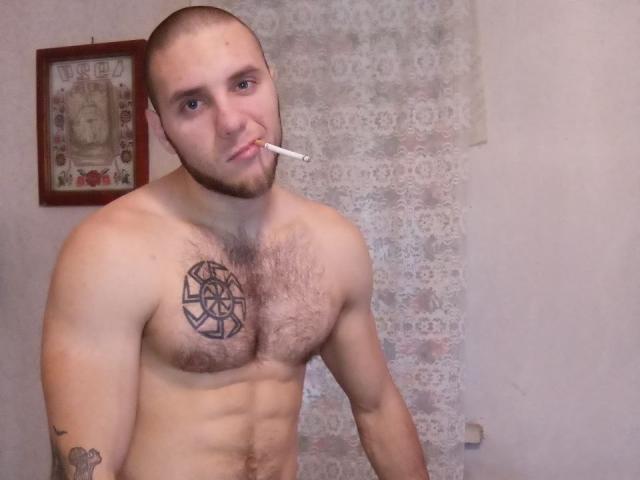 Shirtless JordanDiary with a cigarette in his mouth, bald with a beard, tattoo on chest, hairy chest, and tattoo on right arm, standing in a room.
