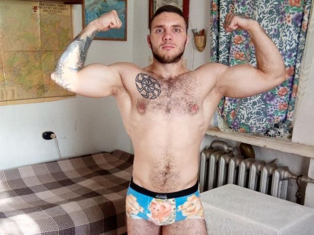 Muscular JordanDiary with tattoos, flexing both biceps, hairy chest, and wearing colorful underwear with a pig face, standing in a room. JordanDiary has a serious expression.
