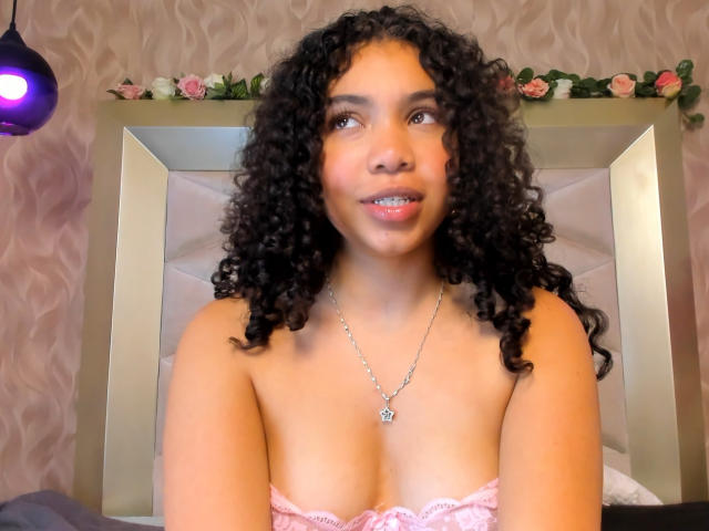 GaiaBlar with curly black hair, light brown skin, and small tits, wearing a pink lace top, necklace with a star pendant, and a gentle smile in a bedroom.