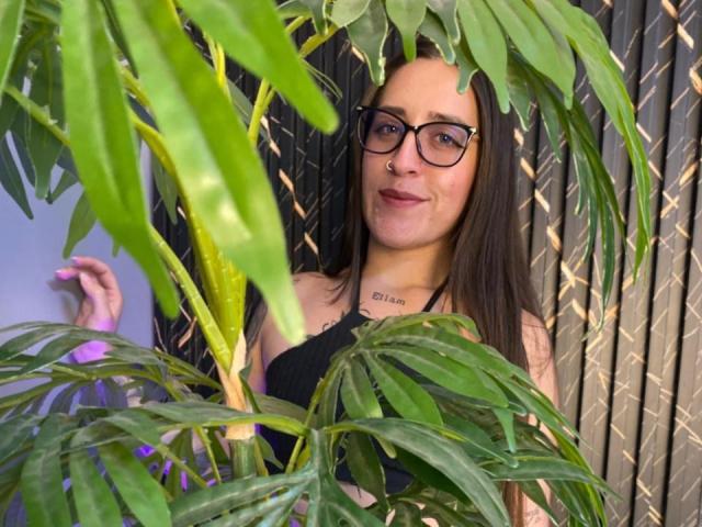 VelvetGreeny with glasses, nose ring, and black top, standing behind green plant leaves, smiling, showing small tits, tattoo on neck, holding plant stem.