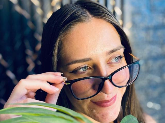 VelvetGreeny with glasses, smiling, holding them with one hand, showing blue eyes, brown hair, and a slight tan, with a green leaf in the foreground.