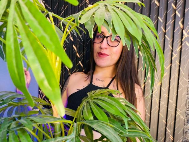 VelvetGreeny with long brown hair, black glasses, and a black top, standing behind green leaves, showing a subtle smile, tattoo on neck, against a dark metal fence.