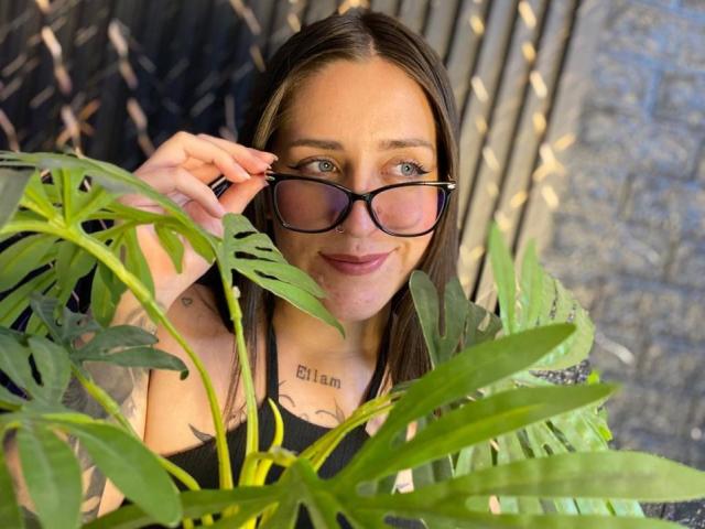 VelvetGreeny with glasses, smiling, brown hair, blue eyes, black tank top, visible tattoo on chest, green plant in foreground, wooden background.