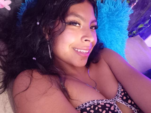 SashaLia with curly black hair, brown skin, smiling, wearing a leopard print bra, blue furry pillow behind her, purple lighting. Boobs visible, eyeliner, long eyelashes, silver earrings.