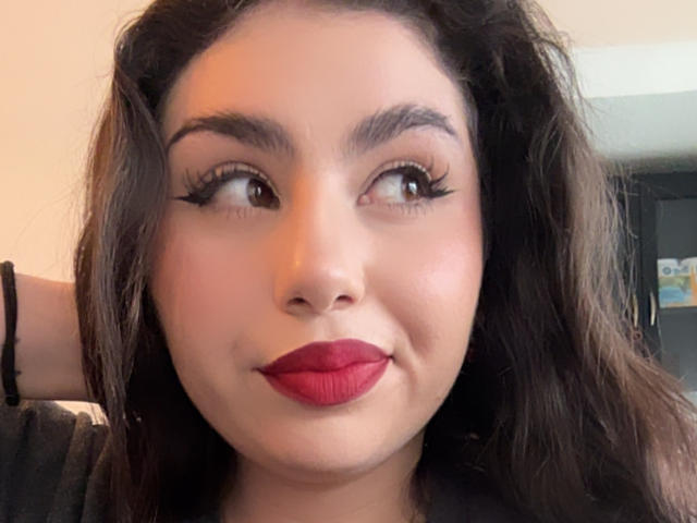 VenusxMusa has dark wavy hair, red lipstick, and thick black eyelashes. She's looking to the side with a subtle smile. Her tan skin is smooth and even.