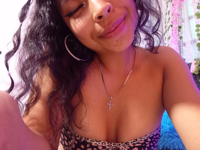 SashaLia with wavy black hair, smiling, wearing hoop earrings and a leopard-print strapless top, showing her tits, wearing a cross necklace, in a pink-lit room.