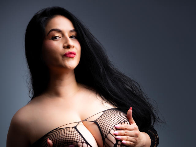 AmbberMoon with long black hair, pink lipstick, and fishnet top covering her large tits, holding one with pink nails, looking slightly to the side.