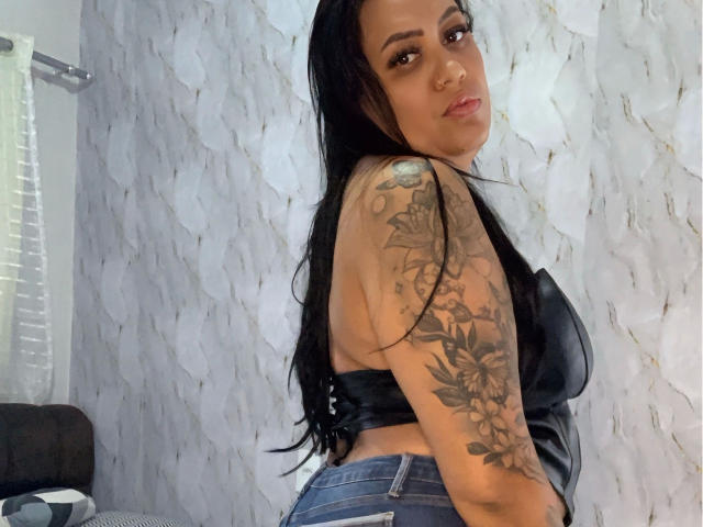 LiraLatinXXX-hot with long black hair, tattoos on right arm, wearing black leather top, blue jeans, looking back with slight smile, nose ring, and eyebrow piercing.