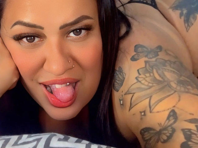 LiraLatinXXX-hot with black hair, brown eyes, nose ring, and tongue out, showing tattoos of butterflies and flowers on her shoulder, and a rose on her upper arm.