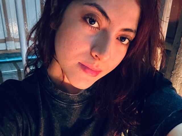 SpencerLe with wavy brown hair, dark eyebrows, and a slight smile, wearing a dark shirt, has a cute face and a small nose. Soft light highlights her skin.