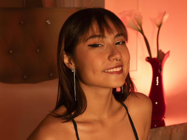SaritaLeone with medium brown skin, straight brown hair, black eyeliner, nose ring, and long earrings, smiling with visible teeth, wearing a black strappy top. Red lighting in background.