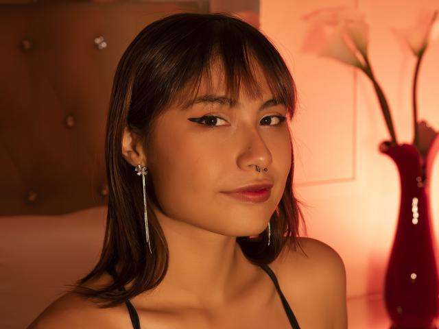 SaritaLeone has medium brown skin, straight black hair with bangs, a nose ring, and black eyeliner. She's wearing a black spaghetti strap top with silver earrings.