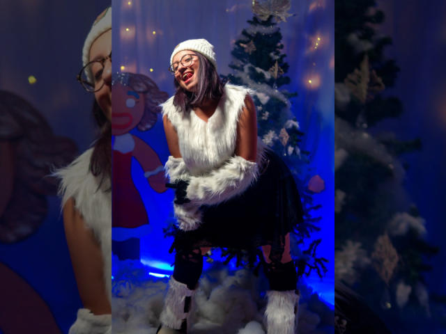 CristinaSmit with white fur vest, black skirt, white fur hat, and boots, bending forward, smiling, wearing glasses, standing in front of a snow-covered Christmas tree.