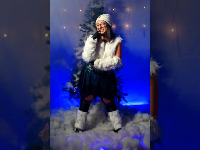 CristinaSmit with white fur vest, black skirt, white boots, and knit hat, standing on fluffy blue snow, smiling, showing her tits under the fur vest. Christmas tree in background.