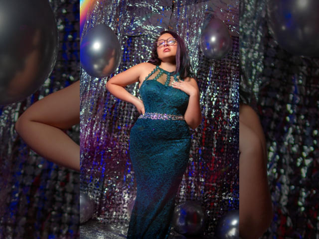 CristinaSmit in a shiny green lace dress, standing with one hand on her hip, wearing glasses, surrounded by silver balloons, her ass slightly visible in the background.