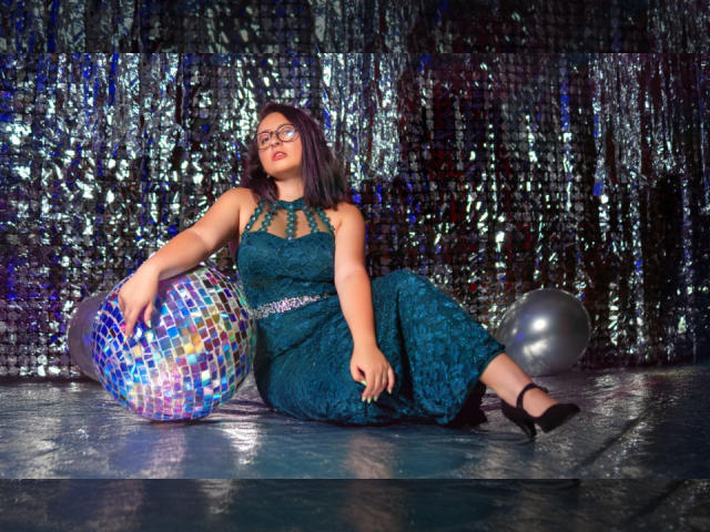 CristinaSmit with glasses, wearing a teal lace dress, sitting on a shiny floor, legs crossed, black heels, holding a colorful mosaic ball, silver balloon nearby.