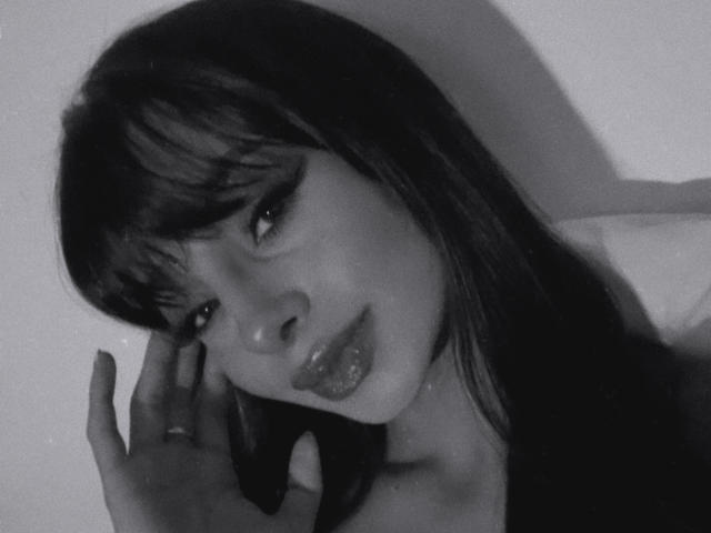 Black and white selfie of ThaliaCollins with dark, straight hair, bangs, and thick eyelashes. She's biting her glossy, shiny lips, touching her face with a ringed finger.