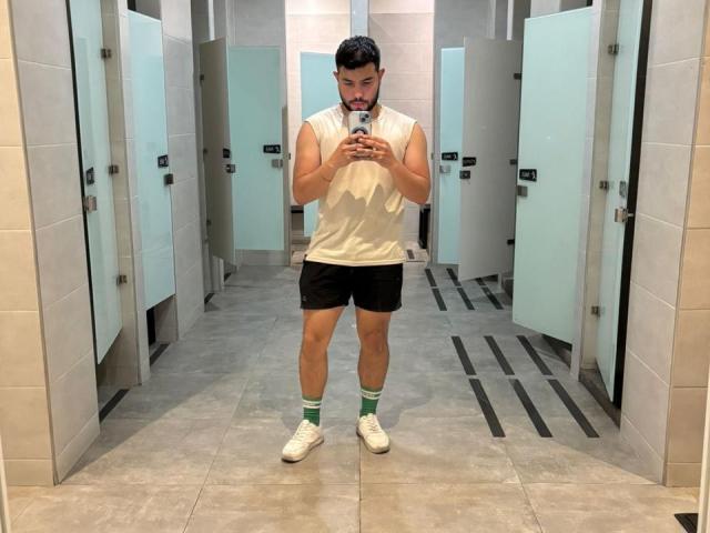 NiganDuquette stands in a bathroom, wearing a sleeveless white shirt, black shorts, green socks, and white sneakers, looking at his phone with a slight beard and black hair.