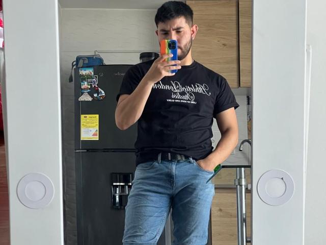 NiganDuquette, standing, taking a selfie, wearing a black t-shirt, blue jeans, with hands in pockets, black-framed mirror, gray refrigerator, wooden cabinets, short black hair, trimmed beard.