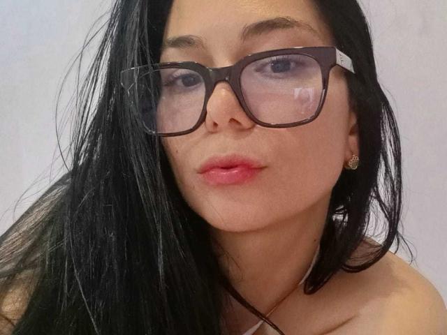 BiancaScott, con gli occhiali neri, le labbra rosa e i lunghi capelli neri, indossa una collana, ha un viso carino ed è rivolta verso la fotocamera con un leggero sbuffo..