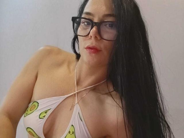 Bianca Scott, con cabello negro largo, usando gafas, y un top blanco de tirantes con estampados de caritas sonrientes verdes, mostrando sus pequeños senos y un collar delgado, sobre un fondo liso..