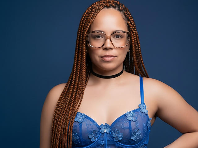 ElennaCruz with long braided brown hair, wearing blue lace bra, glasses, black choker, looking straight at camera, has small tits, medium skin, and a confident expression.