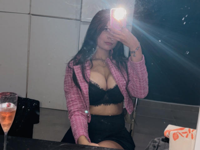 KellyCostaX with big tits in black lace bra, pink cardigan, and black skirt, taking mirror selfie, holding phone, with glass of pink drink on table.