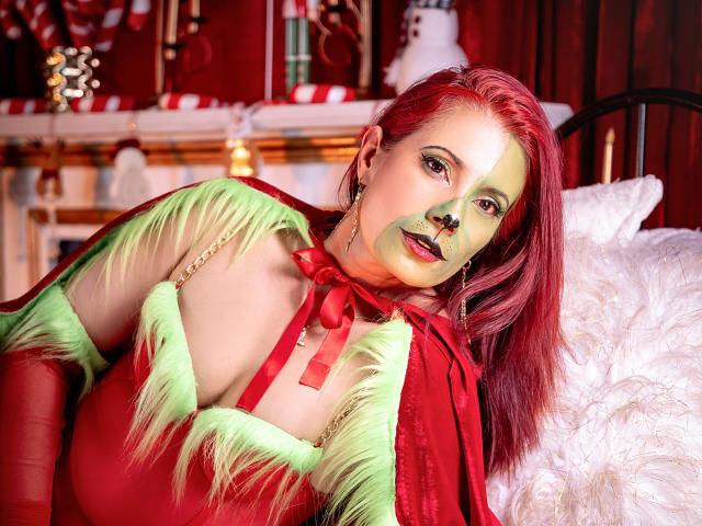 AdaMclain with red makeup, green and red feathered dress, red bow, and green face paint, leaning on white pillow, red hair, looking at camera, wearing red gloves.