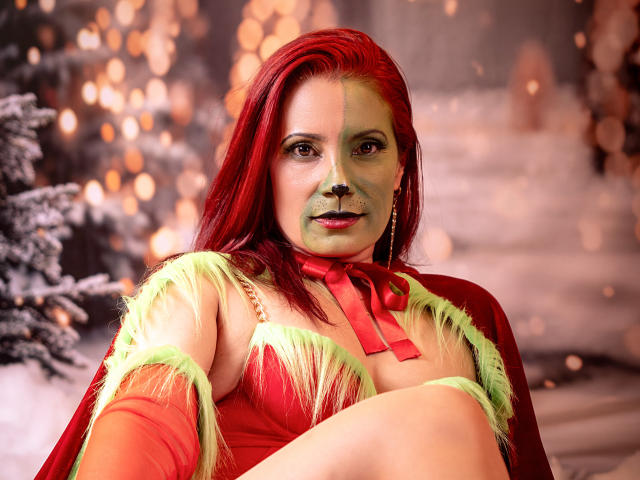 AdaMclain con i capelli rossi, il viso dipinto di verde, una parte superiore decorata con piume rosse e verdi, un nastro rosso e degli orecchini, che mette in mostra il seno e un carino naso nero, in un contesto festoso..