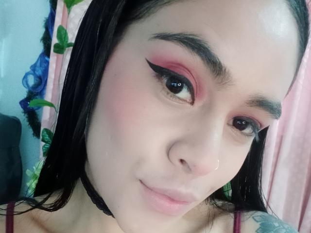StellaBeckker with pink eyeshadow, black eyeliner, and black eyebrows, wearing a black choker, has a small nose ring, and a tattoo on her right arm.