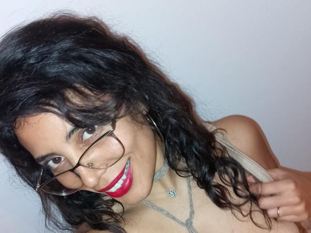 AdamasMoon with curly black hair, glasses, nose ring, red lipstick, necklace, and hoop earrings, smiling at the camera, holding a strap. Her tits are slightly visible.