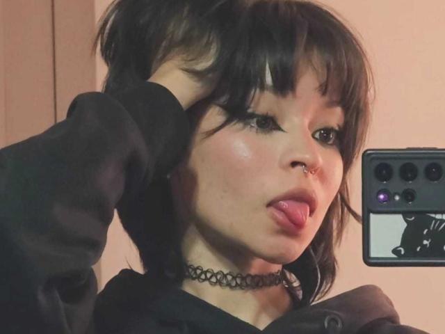 YaikuRb has dark brown hair, a nose ring, and is sticking out her tongue with her tongue slightly out. She's wearing a black choker and a black hoodie.