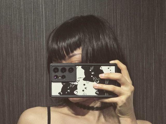 YaikuRb with black bob, holding phone covering face, black choker, white-black phone case with cat faces, taking mirror selfie, visible shoulder strap, dark textured wall background.