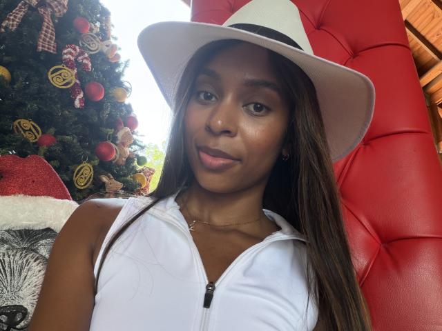 EmmaLatin with dark brown skin, long brown hair, wearing a white hat and sleeveless white top, smiling slightly, red leather chair behind her, Christmas tree with red ornaments on left.
