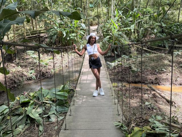 EmmaLatin with medium tits, black shorts, white tank, sunhat, and white sneakers, standing on a wooden suspension bridge, holding ropes, in a lush green jungle.
