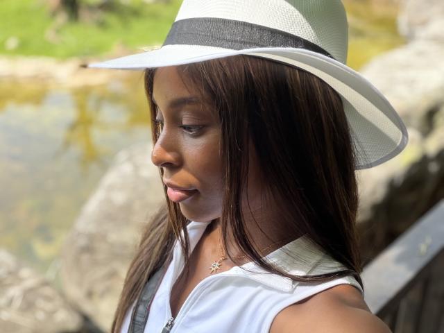 EmmaLatin with dark brown skin, wearing a white sleeveless top, white sun hat with black band, and silver necklace, looking down, brown hair, sunny outdoor setting.