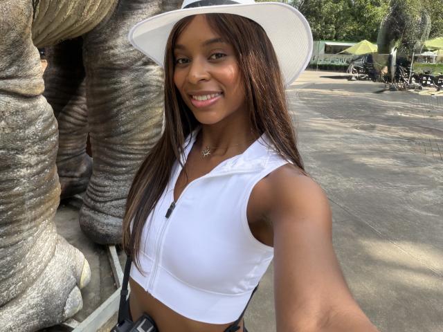 EmmaLatin with brown skin, long straight brown hair, white sleeveless crop top, white hat, smiling, showing small tits, taking selfie in front of large stone sculpture.