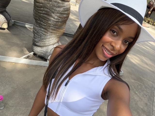 EmmaLatin with dark skin, long brown hair, white hat, white sports bra showing small tits, smiling, standing near a palm tree. Bottle partially visible on left.