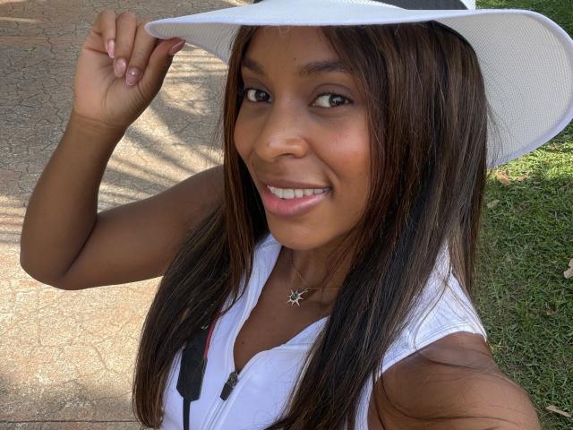 EmmaLatin with brown skin, long straight brown hair, white sleeveless top, white sunhat, star necklace, smiling, holding hat, visible breasts, grass and pathway in background.