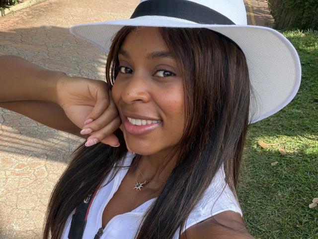 EmmaLatin with brown skin, long straight black hair, white hat, white top, smiling, hand on cheek, grass and pathway in background, wearing star necklace, looking cute.