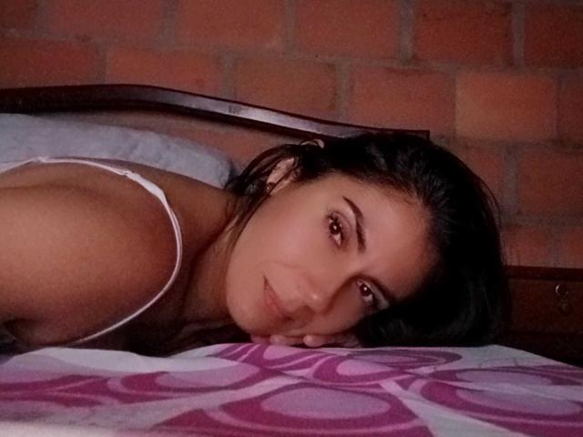 TeacherSexy with dark hair, lying on a bed, head resting on purple-patterned pillow, wearing white spaghetti strap top, smiling softly, brown eyes, brick wall in background.