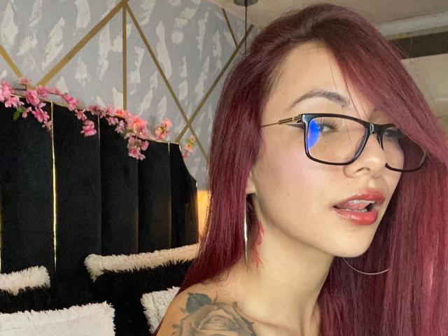 Valery Harllow, com cabelo ruivo, óculos e lábios rosados, tem uma tatuagem no ombro direito e está exibindo seu bumbum pequeno e adorável na foto..