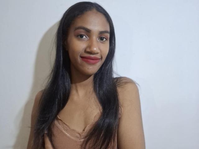 SarahJone has long black hair, brown eyes, and is smiling with red lipstick. She's wearing a lace-trimmed, brown tank top, showing her small tits.
