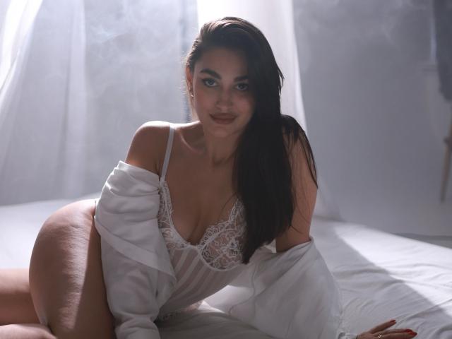 BrunetteBabeXX-hot with long dark hair, white lace lingerie, and off-shoulder shirt, showing big boobs and curvy ass, lying on bed, looking at camera with a soft smile.