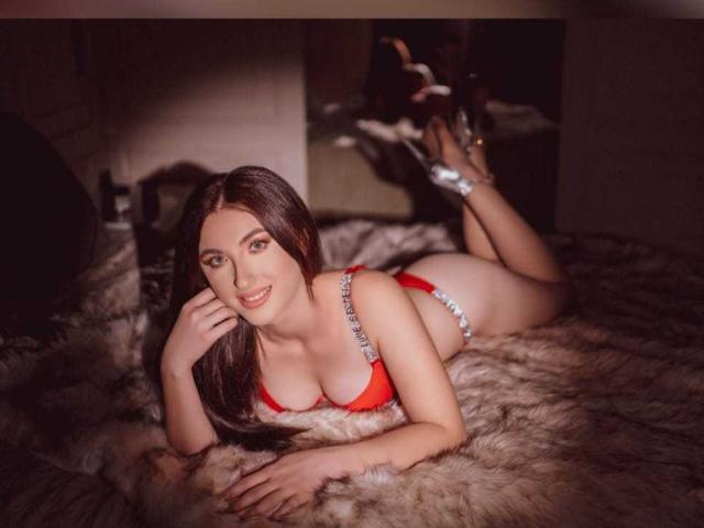 SucreVixen lying on a furry rug, wearing a red bra and thong with silver trim, smiling at the camera, her ass up, legs bent, and brown hair flowing.