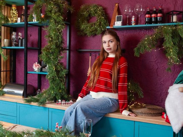 LaraPika with long brown hair, red striped sweater, blue jeans, sitting on a teal cabinet, surrounded by Christmas decor, green wreaths, and wine glasses. Her ass is cute.