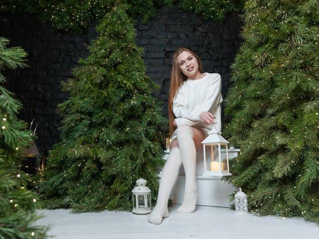 LaraPika with long brown hair, white sweater, and white knee socks, sitting between green Christmas trees, legs crossed, smiling, lanterns with candles on white floor, dark background.